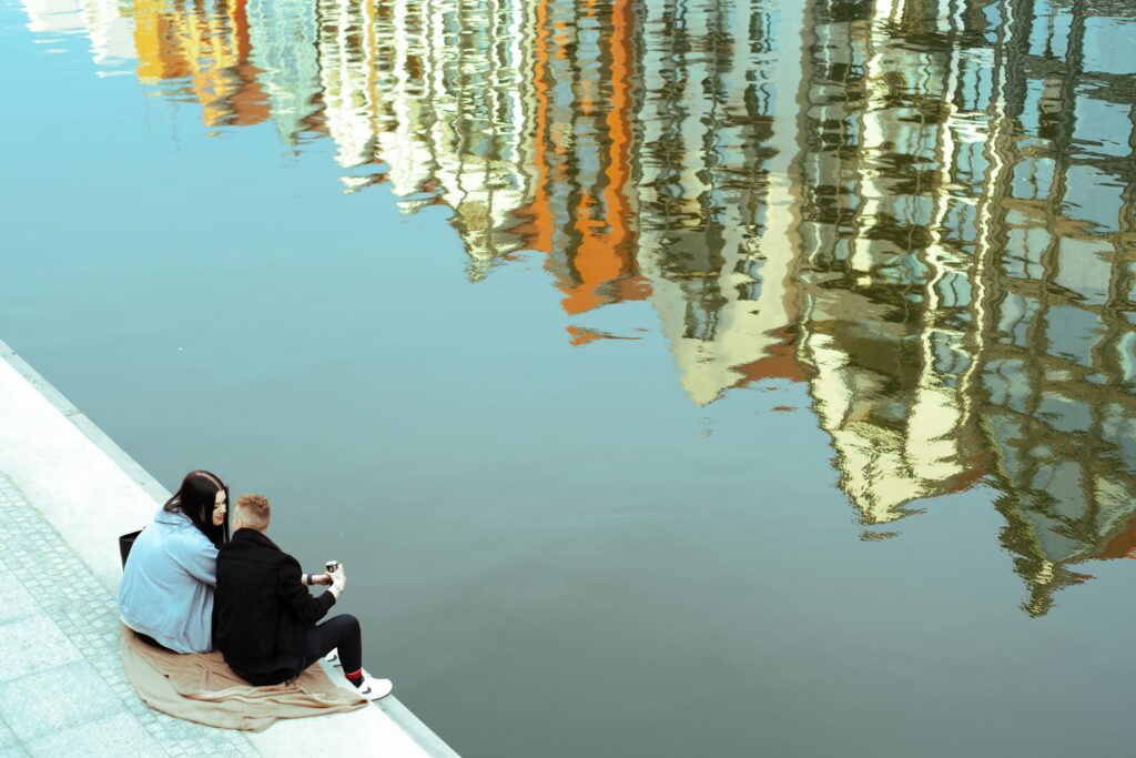 A couple enjoys a peaceful moment by the river in Gdańsk with building reflections in the water.