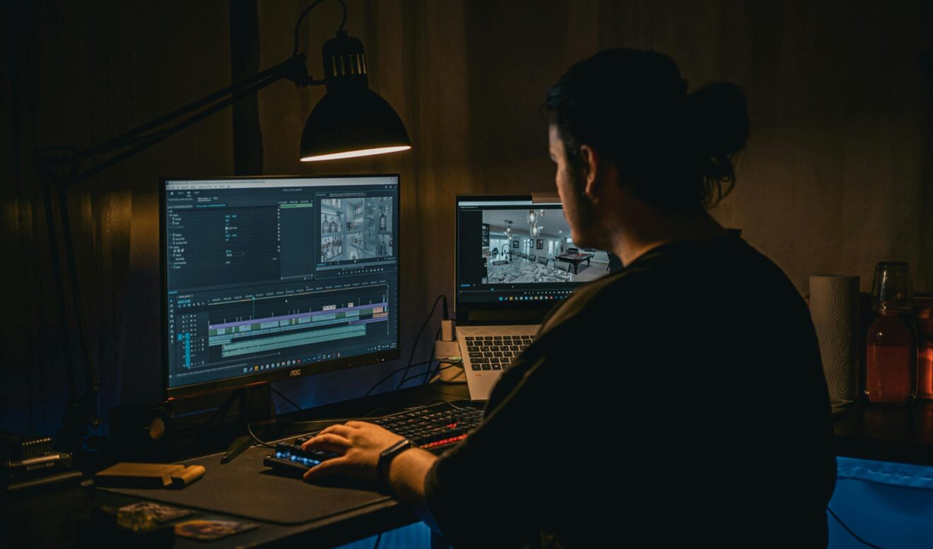 A man works at a computer editing video footage in a dimly lit workspace.