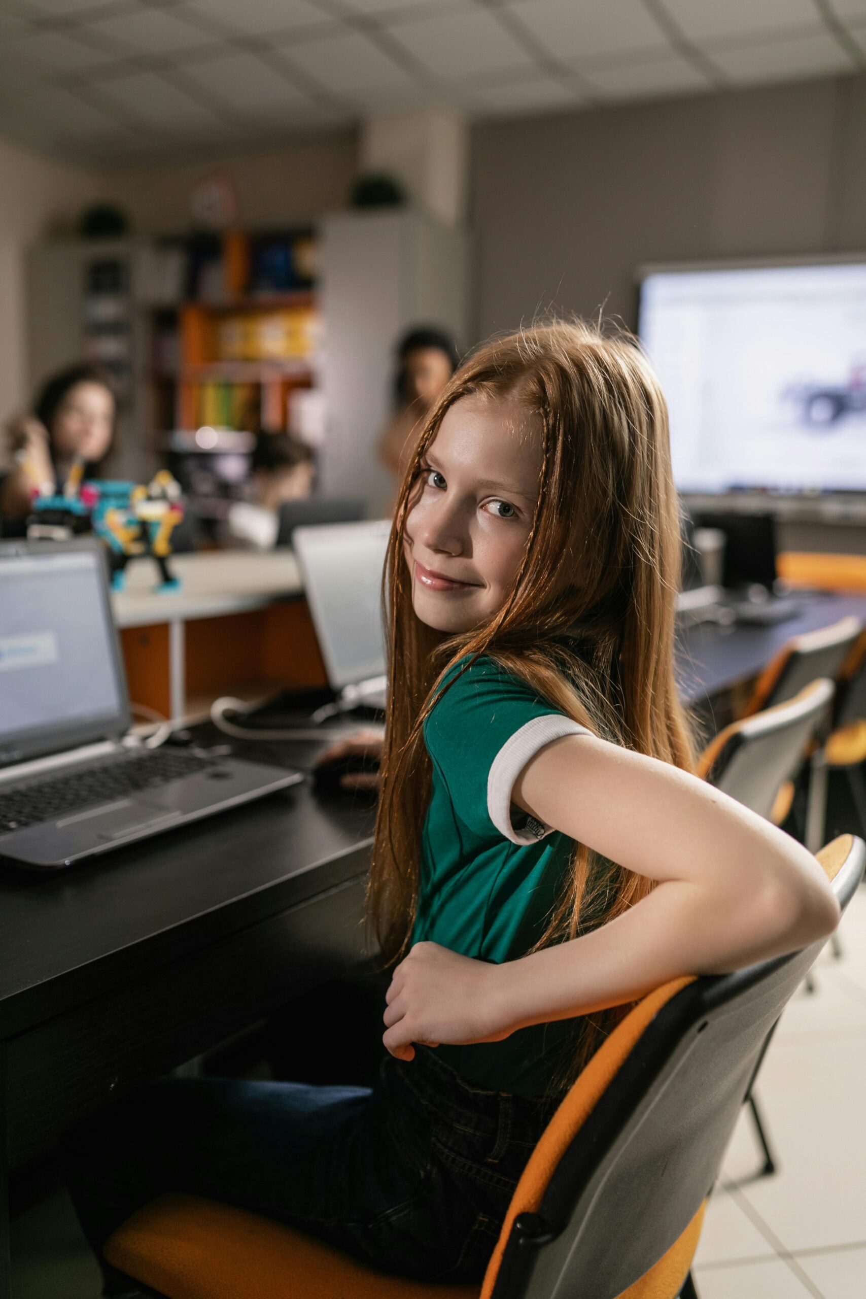 Teen girl smiling at camera while sitting at desk in modern tech classroom.
