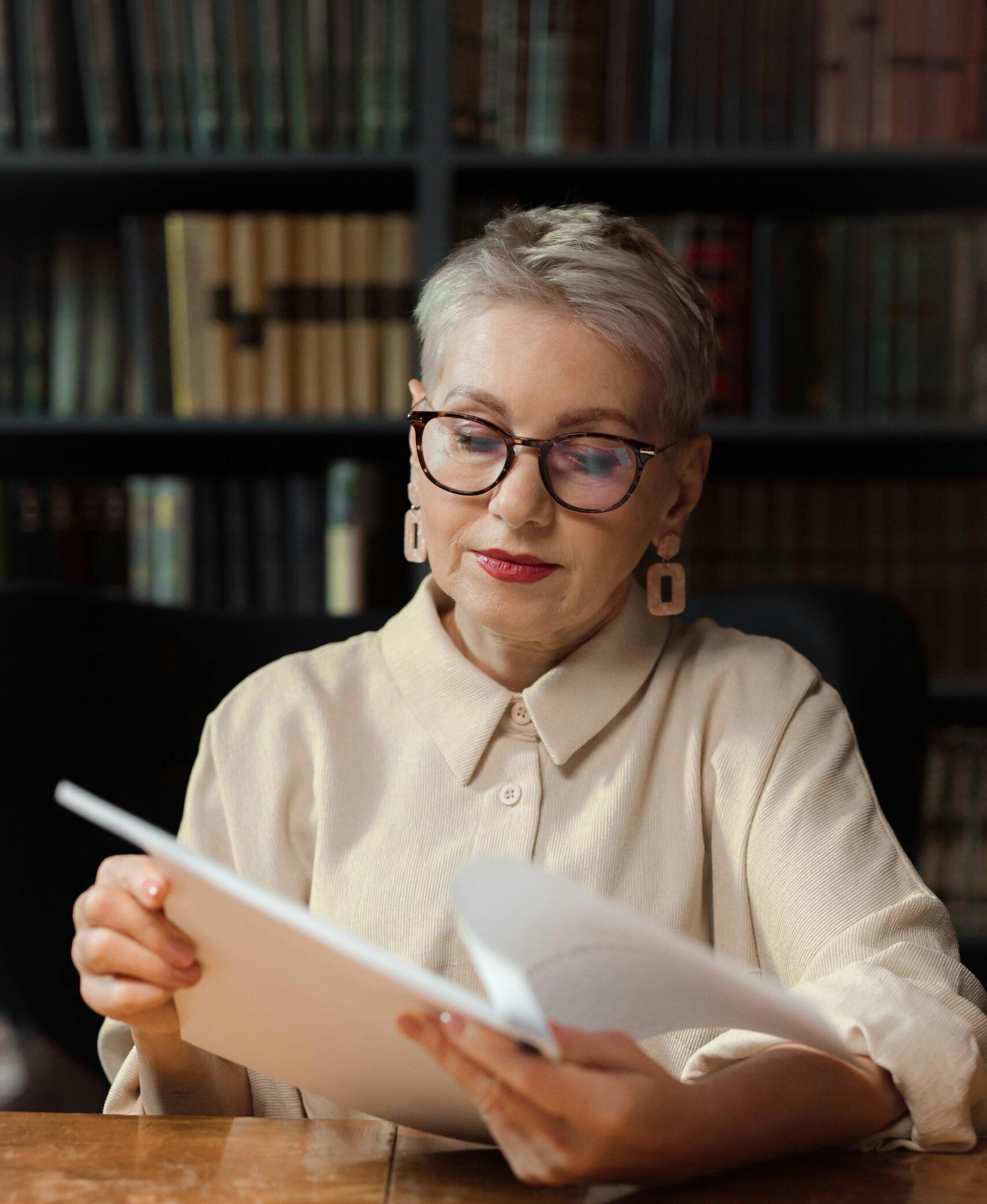 Senior woman reading a script at a wooden table in a cozy library setting.