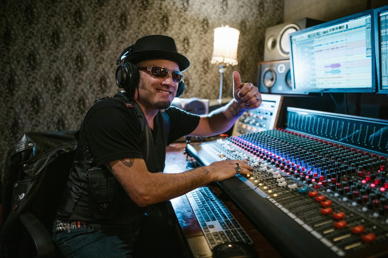 Smiling music producer wearing headphones and sunglasses at a mixing console in a recording studio.