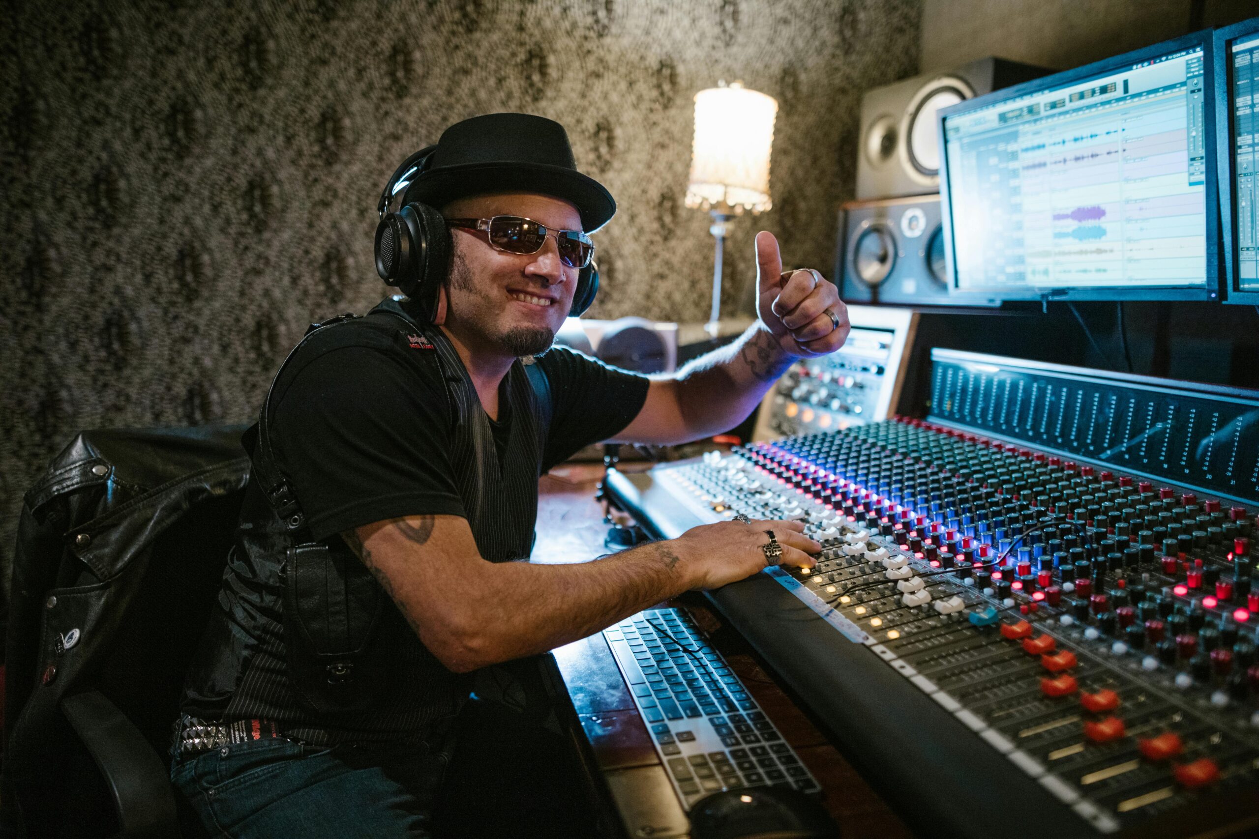 Smiling music producer wearing headphones and sunglasses at a mixing console in a recording studio.